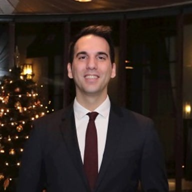 Man in a dark suit and burgundy tie smiles at the camera, standing indoors beside a lit Christmas tree.